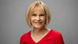 older white woman with blonde bob cut with bangs in a red v neck sweater against a gray background