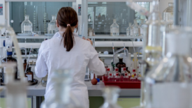 backside view of a chemist in a lab coat working at a table full of glassware