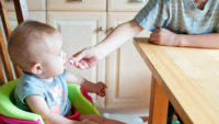 older child feeding snack to baby in high chair