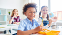 three kids smiling at camera eating lunch at school