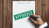 white piece of paper on clipboard with green stamp that says approved and a hand in business suit holding pen in foreground