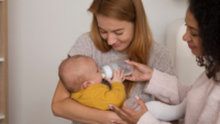 two women feeding bottle of formula to infant